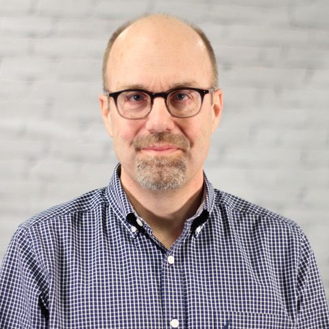 A man wearing glasses and a plaid shirt is standing in front of a brick wall.