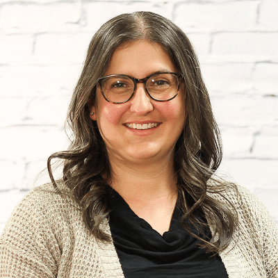 A woman wearing glasses and a sweater is smiling in front of a white brick wall.