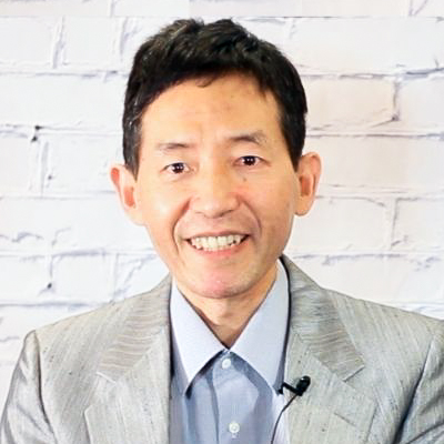 A man in a suit and tie is smiling in front of a white brick wall.