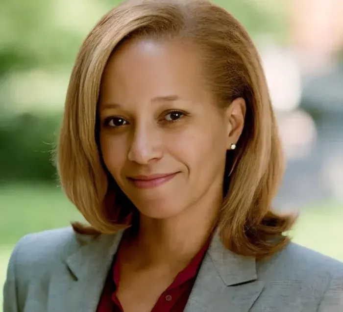 Woman in blazer smiles, looking at the camera outdoors, with a blurred background.