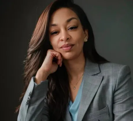 Woman in a grey blazer, resting chin on hand, looking at the viewer.