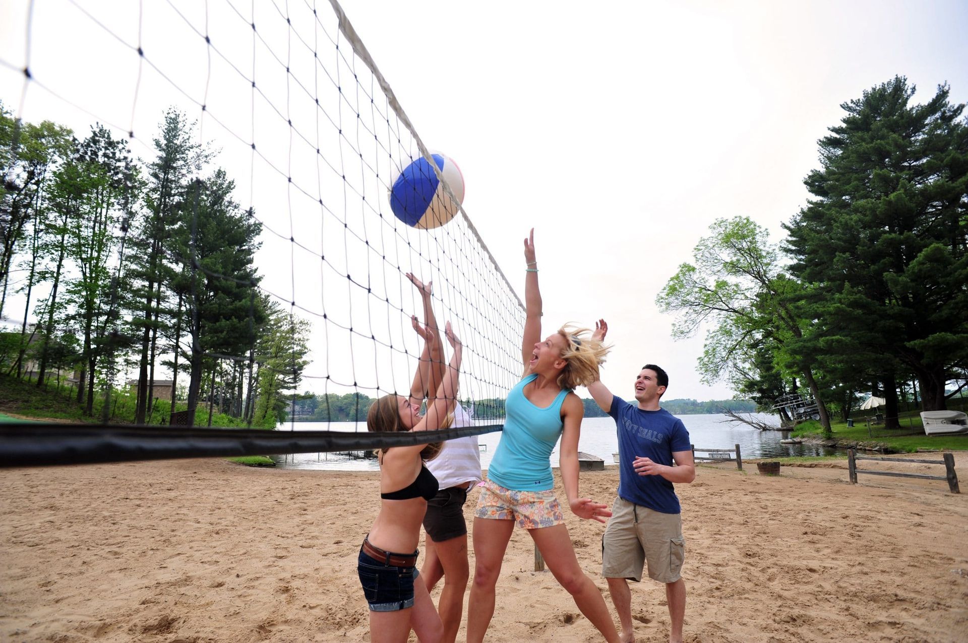 People playing volleyball on a sandy beach near a lake, reaching for a blue and white ball.