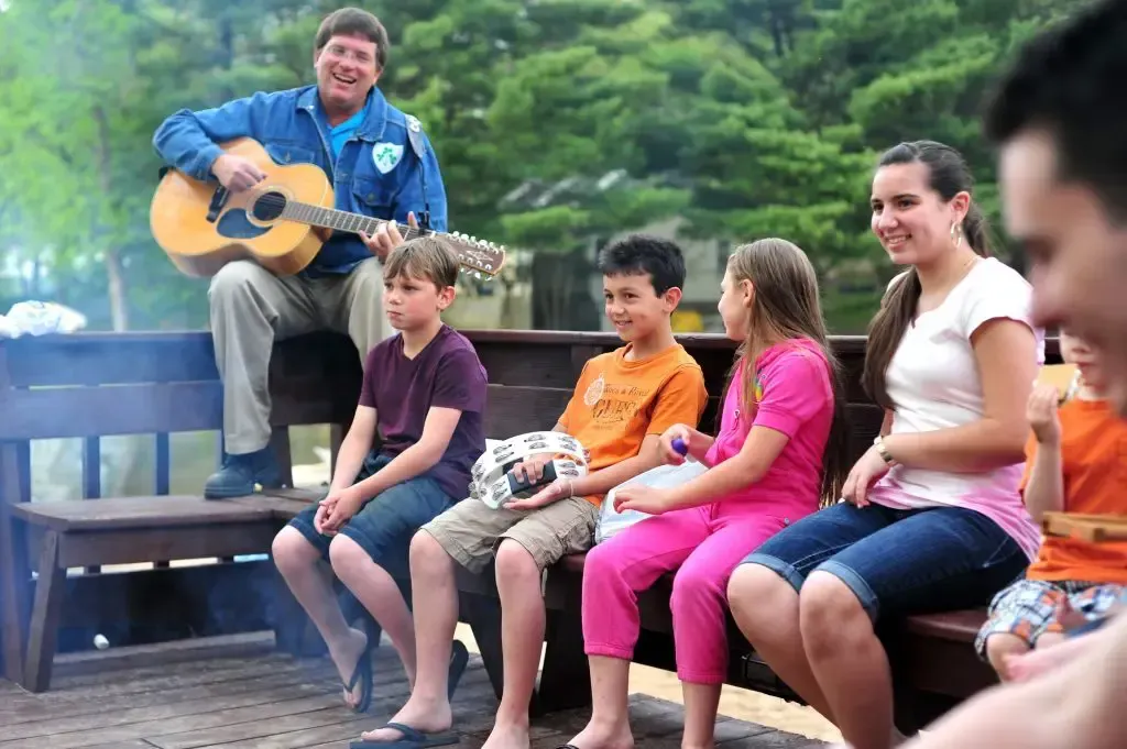 Man playing guitar for a group of children seated on a wooden bench outdoors.