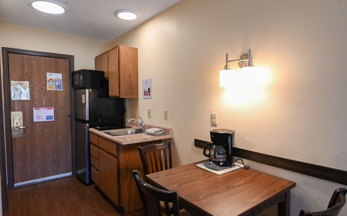 Small hotel room kitchen with microwave, refrigerator, sink, cabinet, table, chairs, and coffee maker.