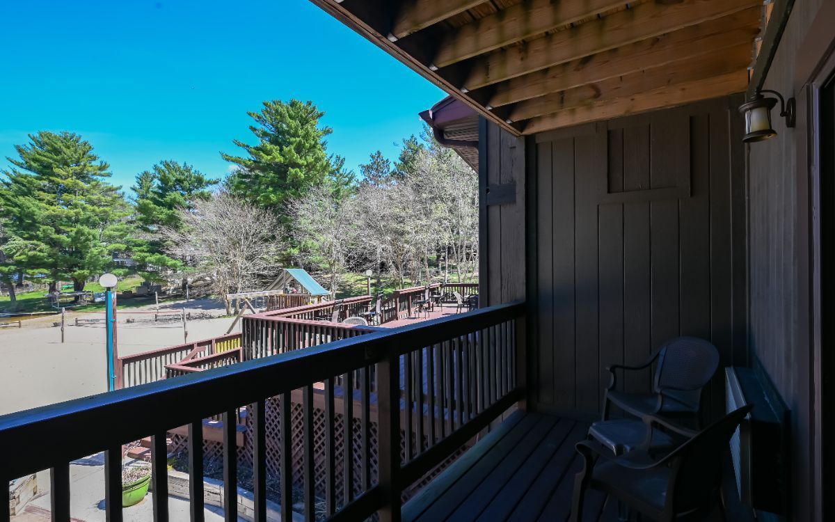 Balcony with dark wood railing, overlooking trees, a deck, and the outdoors; a chair sits on the balcony.