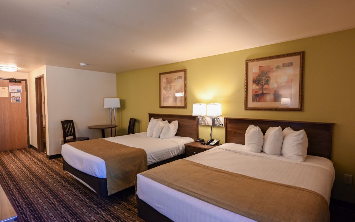 Hotel room with two double beds, nightstand, lamps, artwork, and a patterned rug.