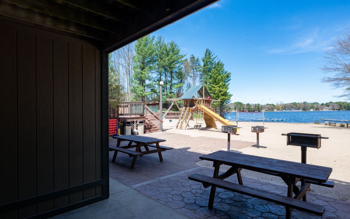Picnic tables and grills on a sandy beach near a playground, with a lake and blue sky in the background.