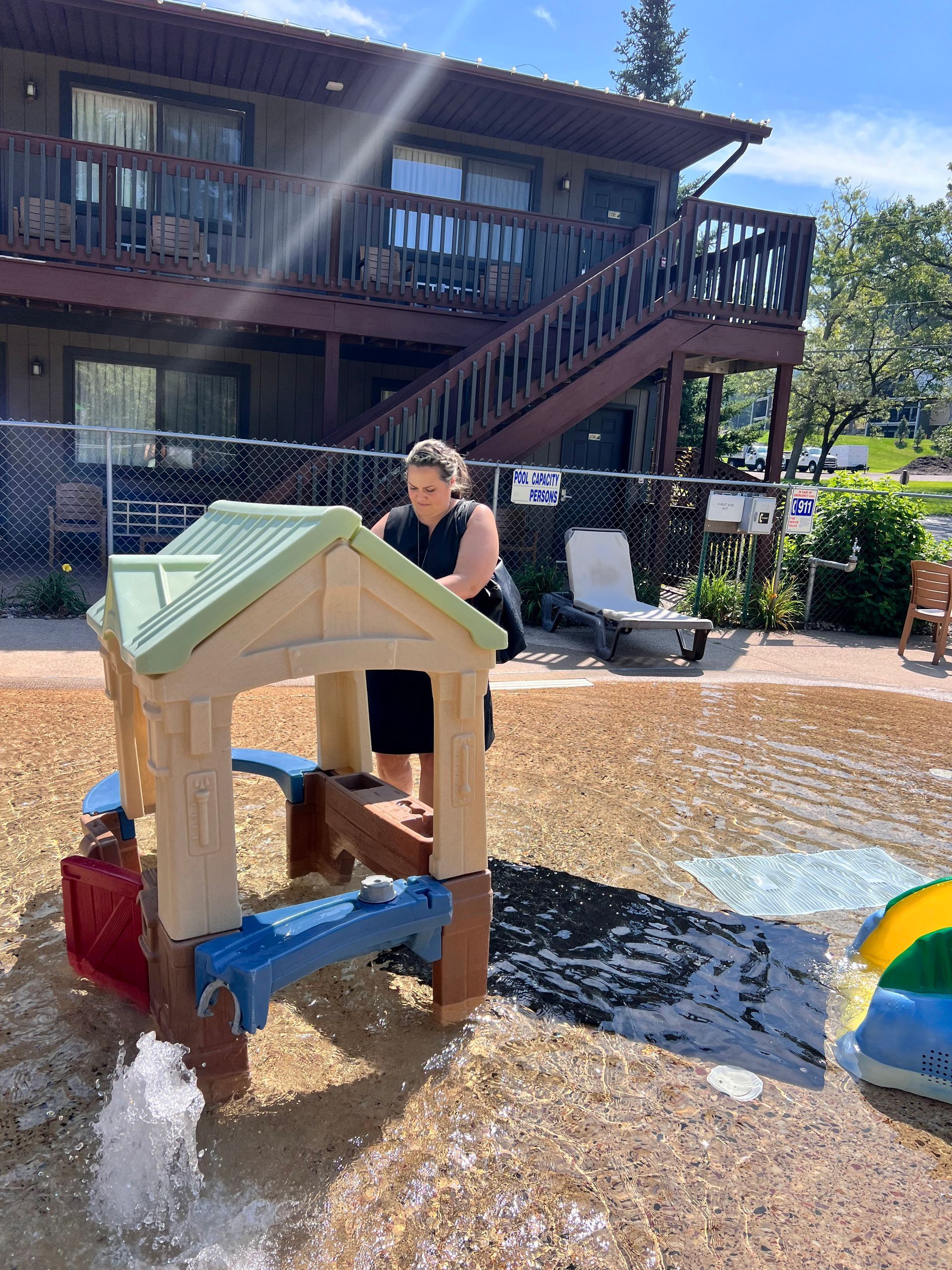 Woman at a children's water play area near a two-story building. Water sprays from the play structure.
