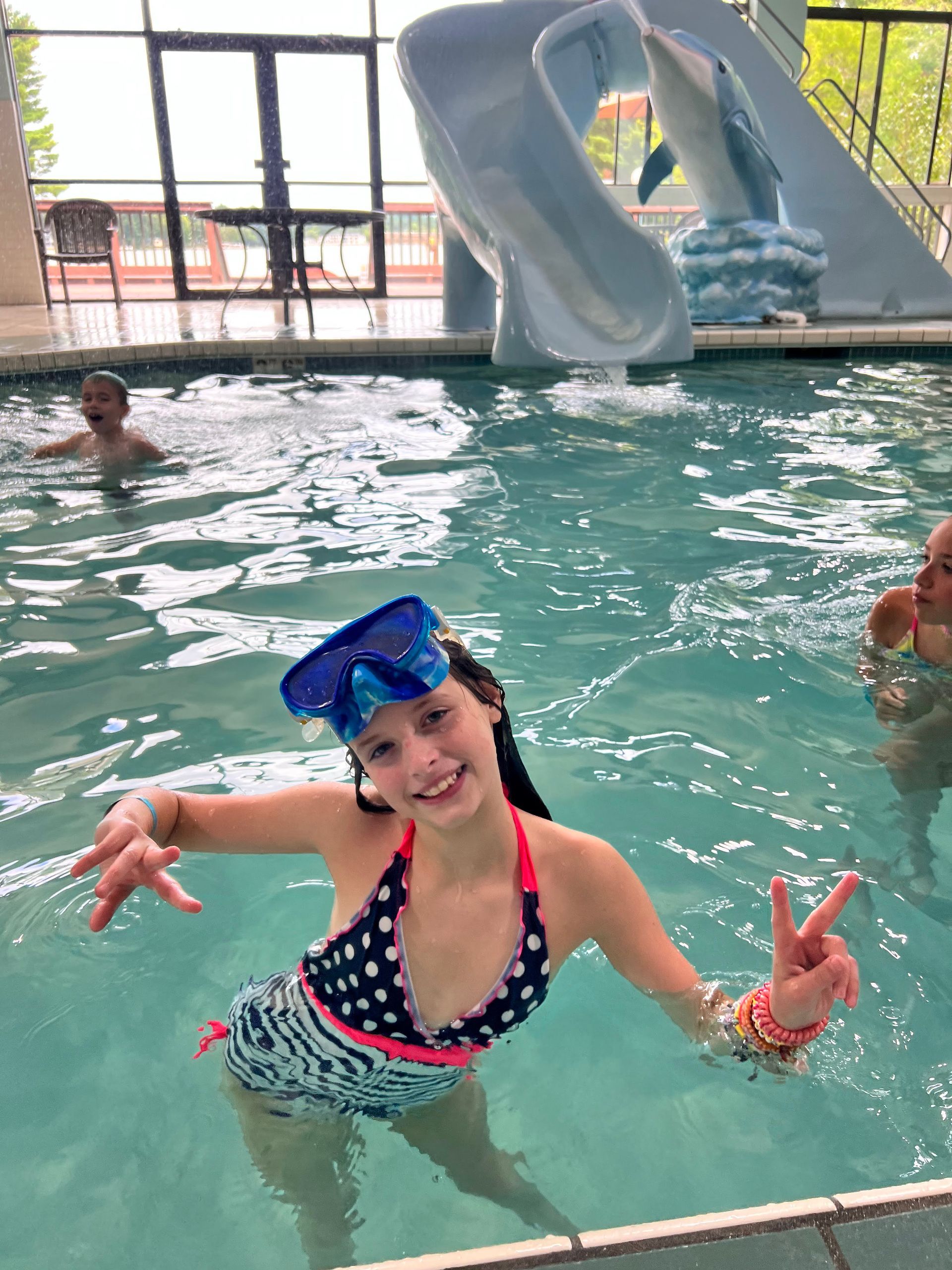 Girl in swimsuit and goggles in indoor pool, making peace signs, smiling. Water slide in background.