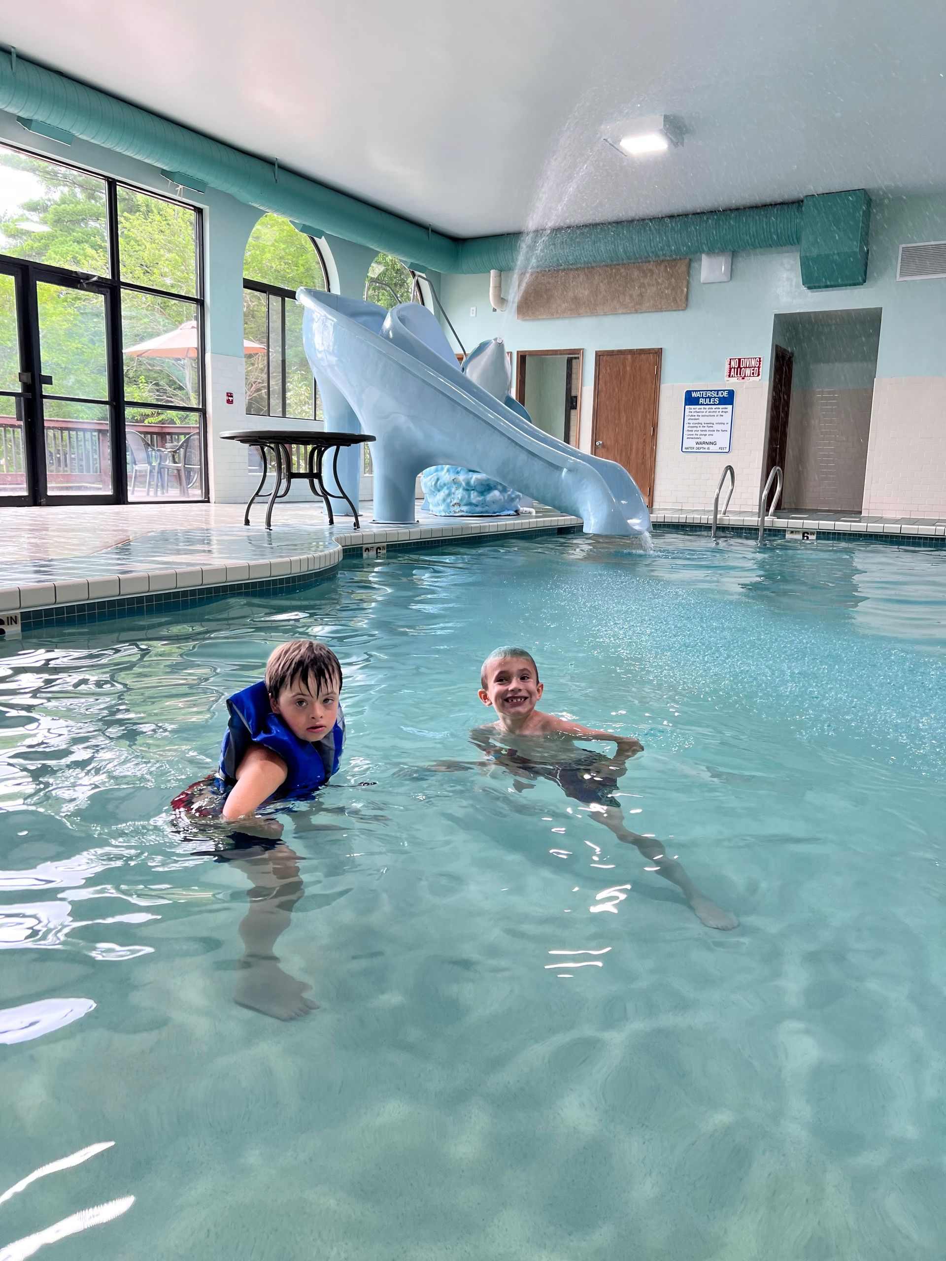 Two boys swim in an indoor pool with a light blue slide and water fountain. One wears a life vest.