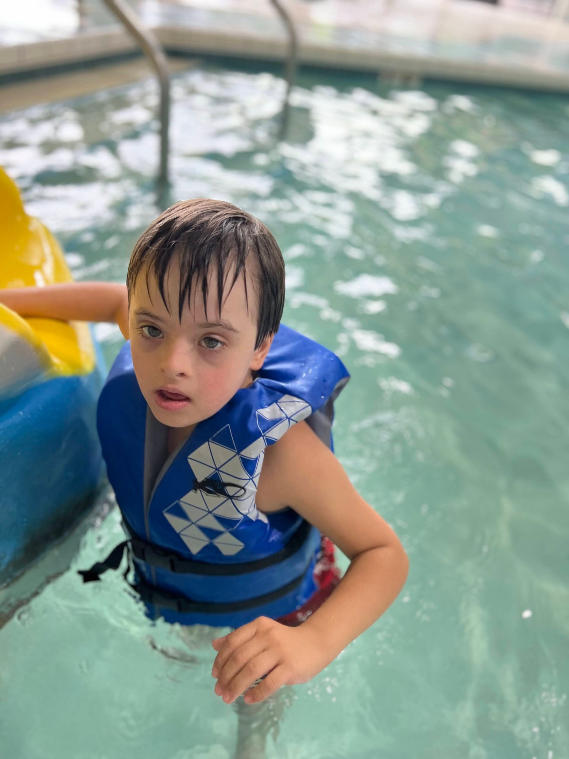 Boy with Down syndrome wearing a blue life vest in a pool, looking at the camera.