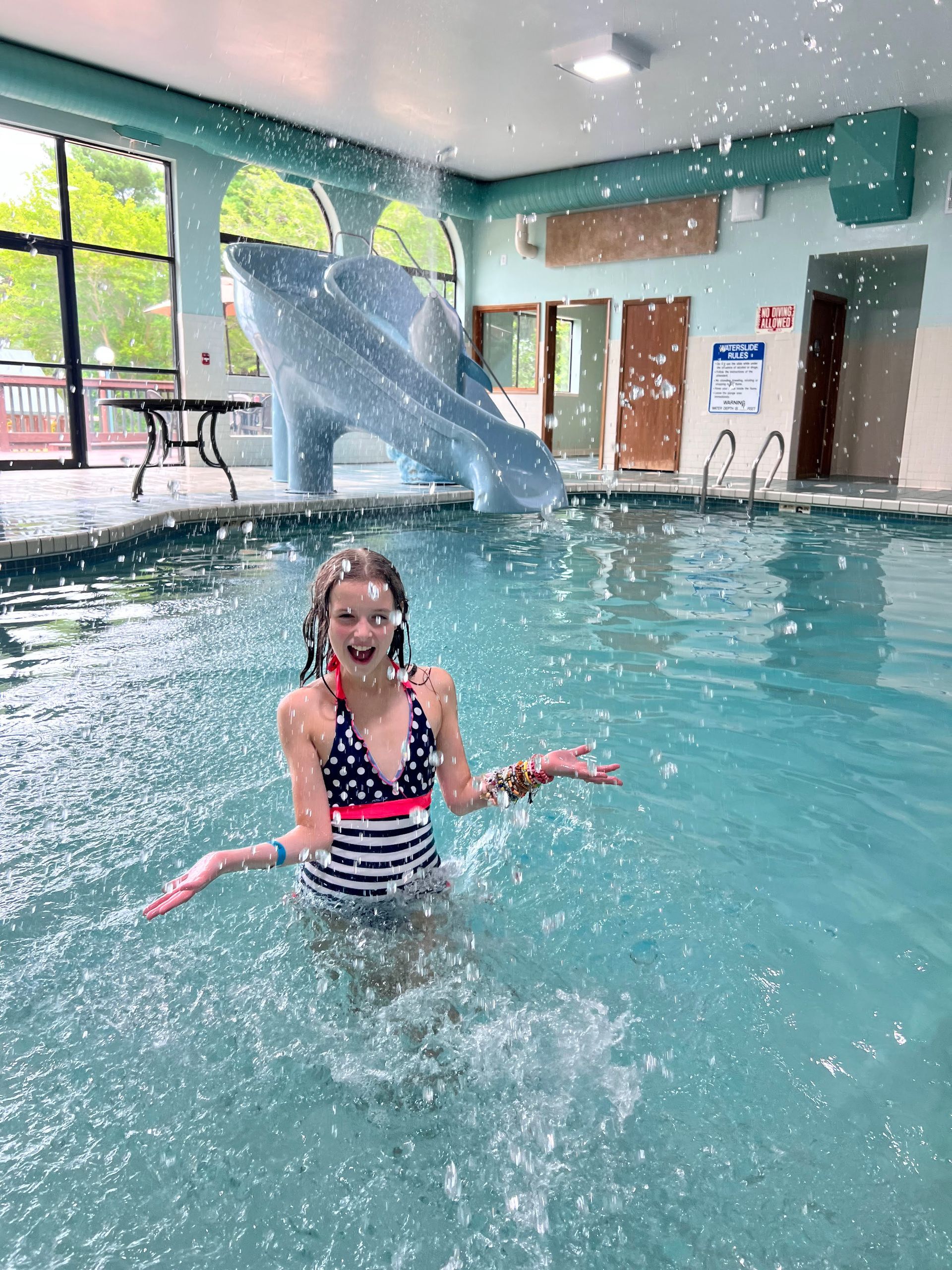 Girl in a swimsuit splashes in a pool with a slide, indoor setting.