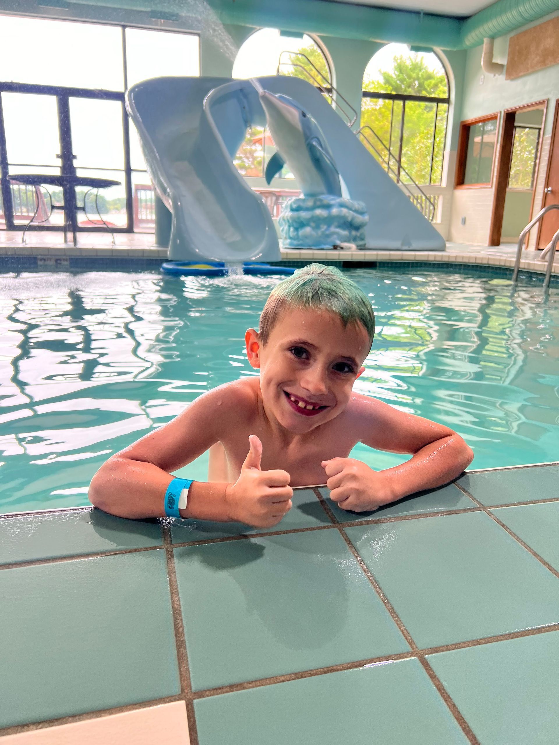 Boy in swimming pool gives thumbs up; blue water slide in background.