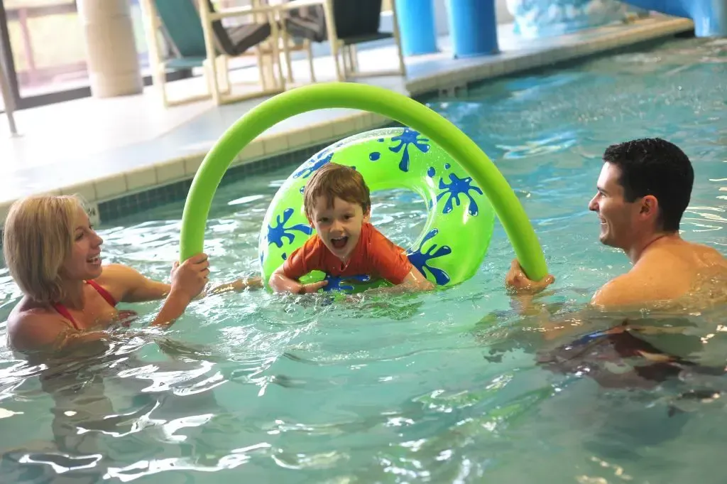 Child in swim ring in pool with adults holding a green pool noodle, all smiling.