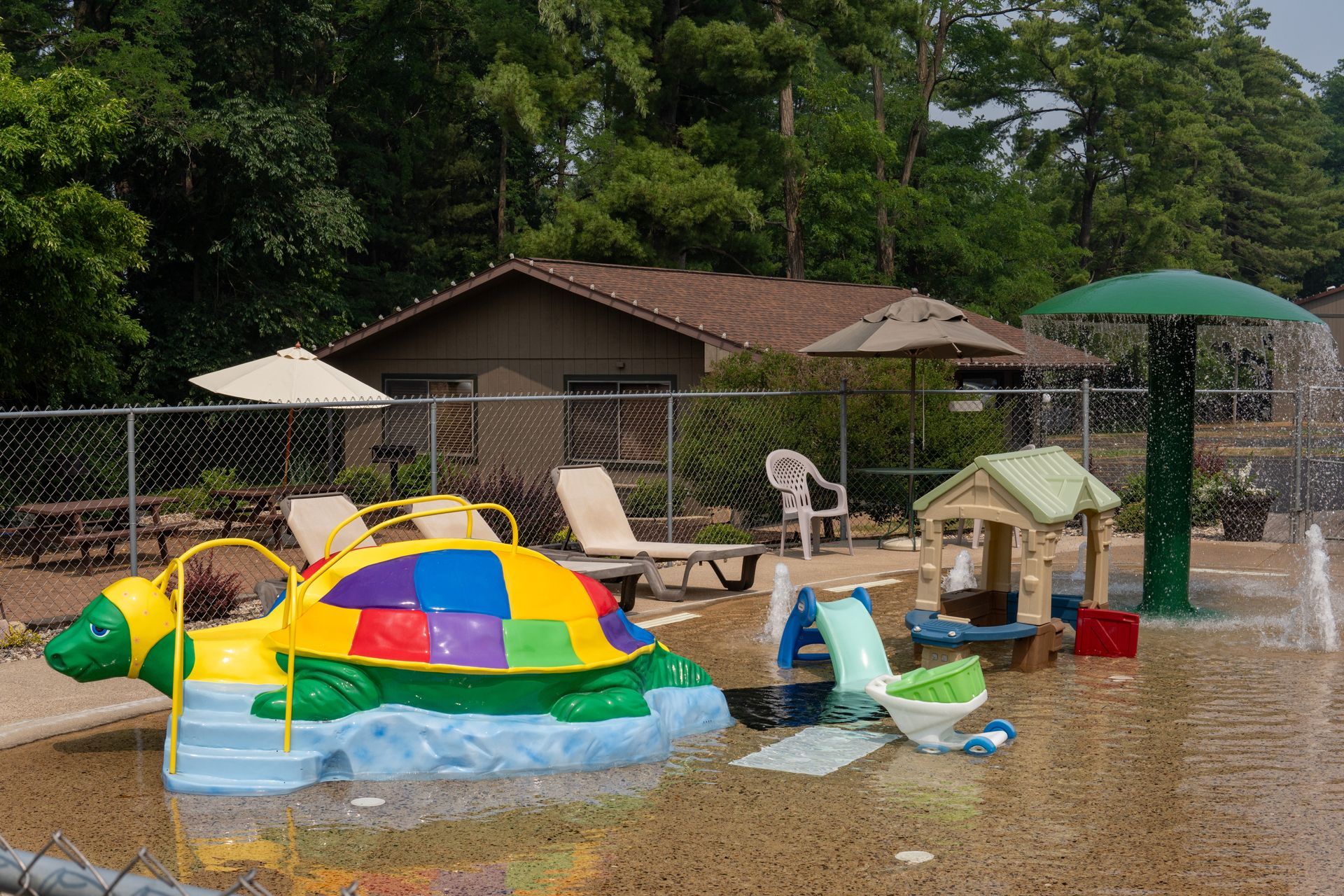 Playground splash pad with turtle, playhouse, and water features near a building.