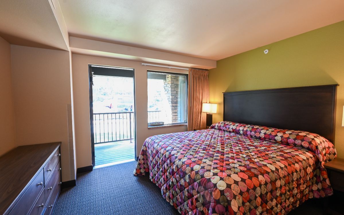 Hotel room with a queen bed, balcony, and dresser. Green wall and colorful bedspread.