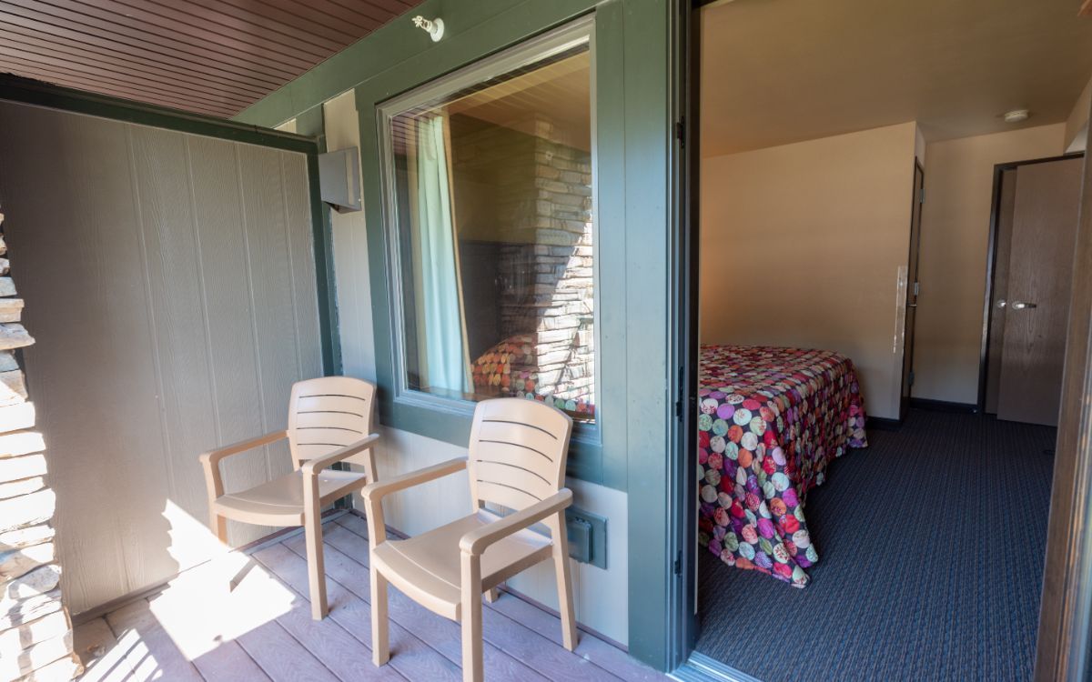 Two chairs on a wooden balcony outside a motel room with a bed and door.