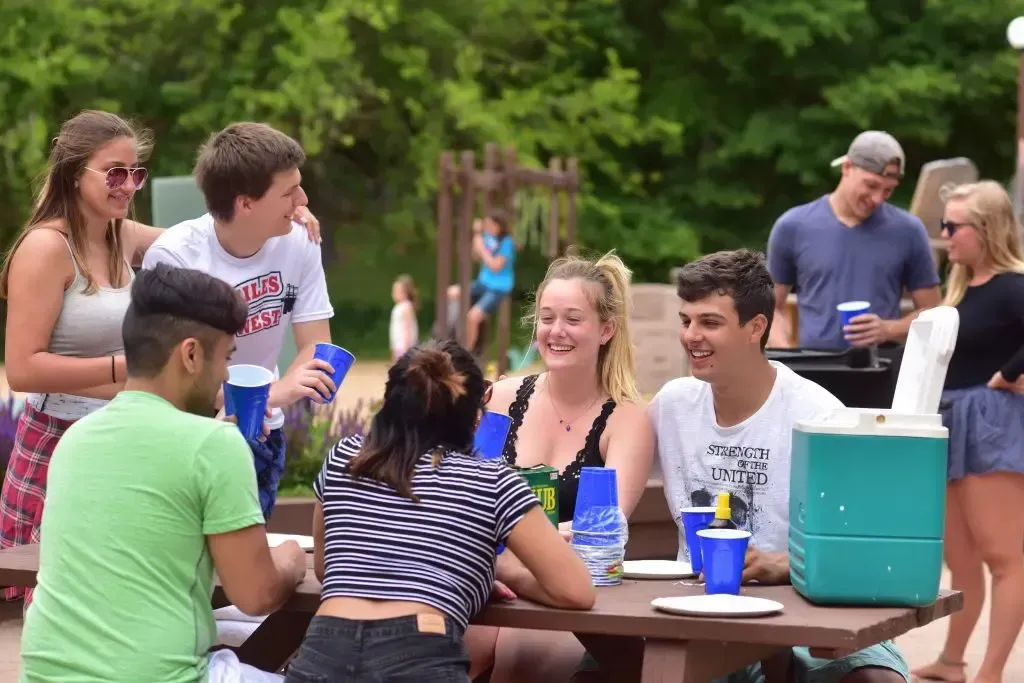 Group of people at a picnic table, smiling and talking outdoors; cooler and cups present.