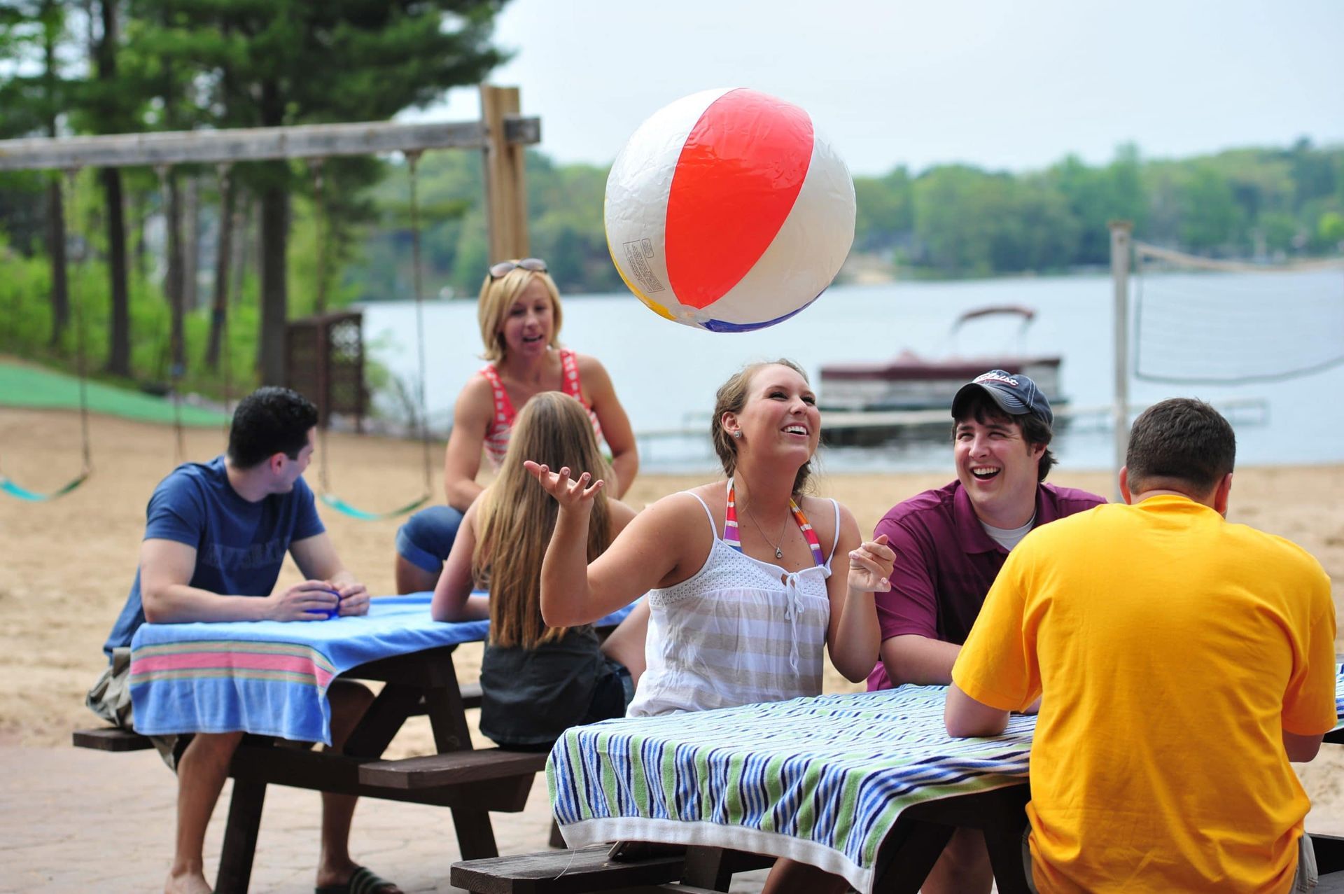 Group plays beach volleyball at picnic tables on a sandy lakeside beach.