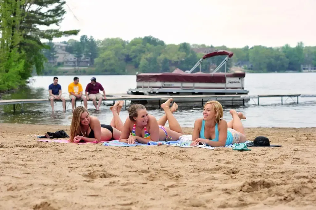 Three women in bikinis sunbathe on a beach, lake and pontoon boat in background.
