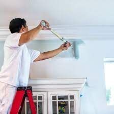 A man is standing on a ladder painting the ceiling of a room.