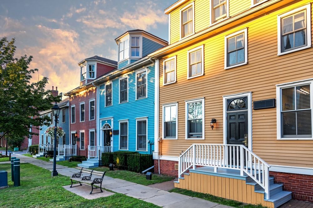 Row of colorful houses with varying colors and architectural styles on a street with benches and a green lawn.