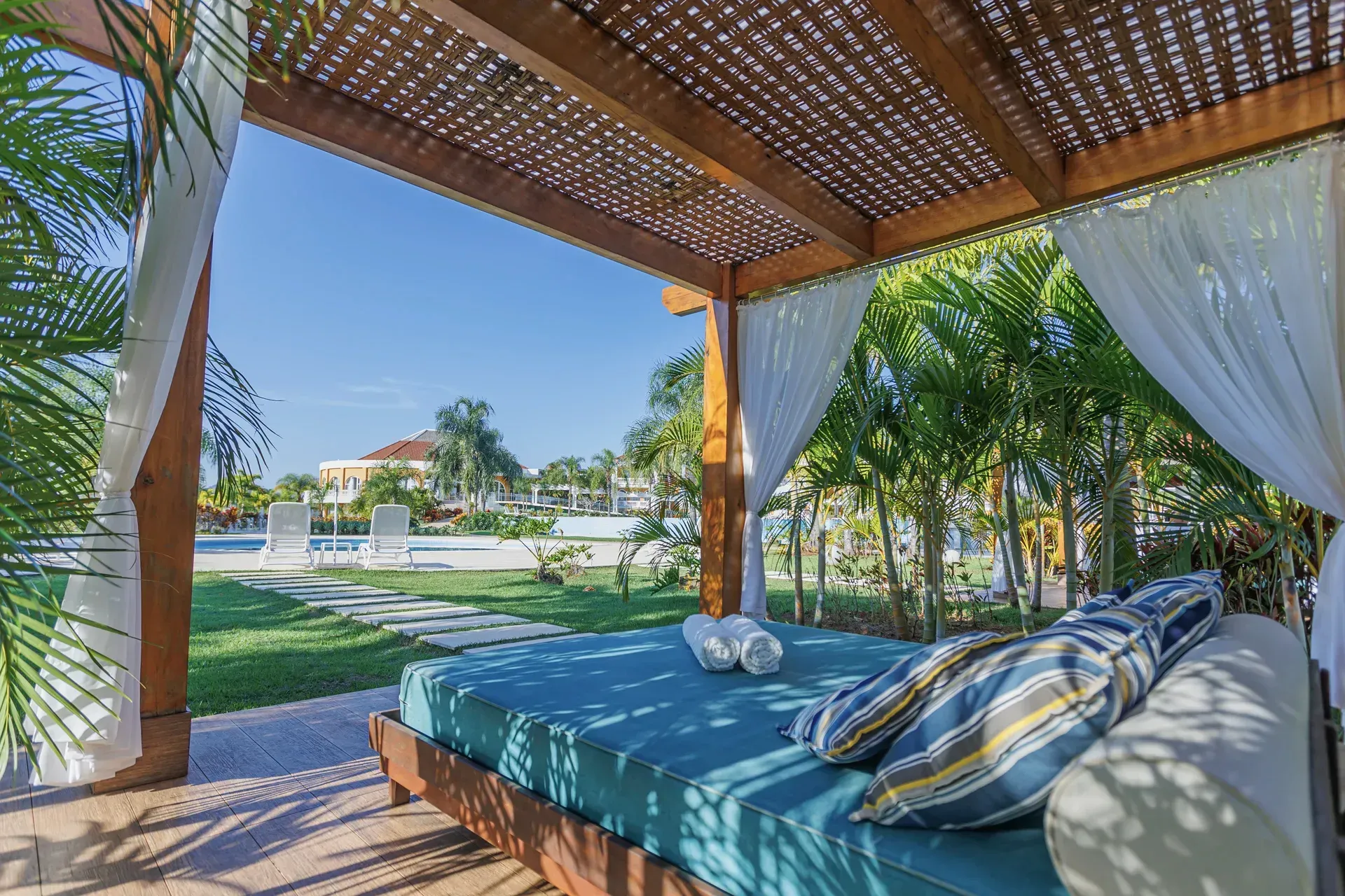 Piscina em um resort tropical com espreguiçadeiras, palmeiras e prédios sob um céu azul.