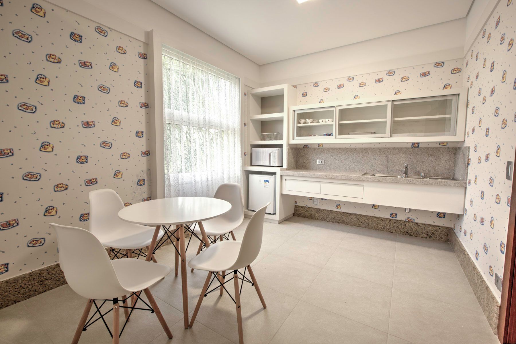 Uma pequena cozinha com mesa e cadeiras brancas, micro-ondas e armários. O papel de parede tem um padrão peculiar.