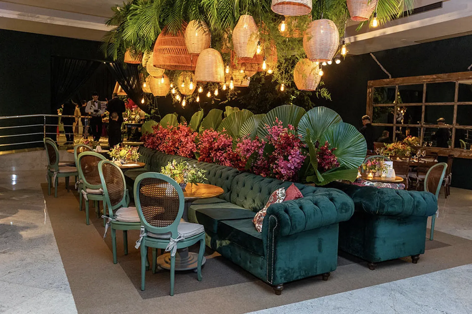 Área de estar verde-esmeralda, arranjos florais exuberantes e lanternas penduradas em um ambiente para eventos.