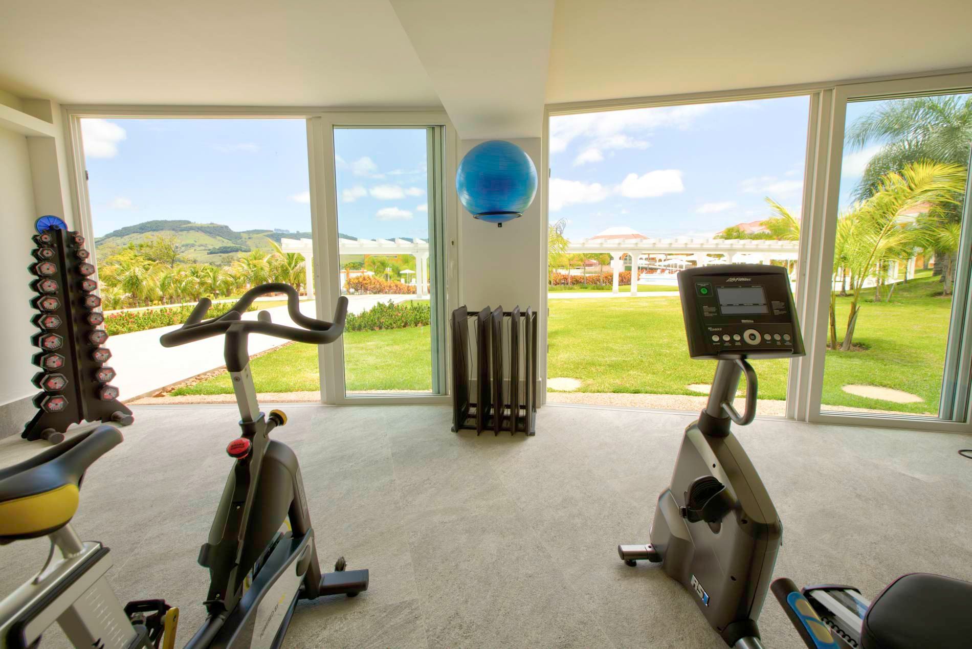 Academia com bicicletas ergométricas, pesos e vista para um gramado e uma paisagem distante através de grandes janelas.