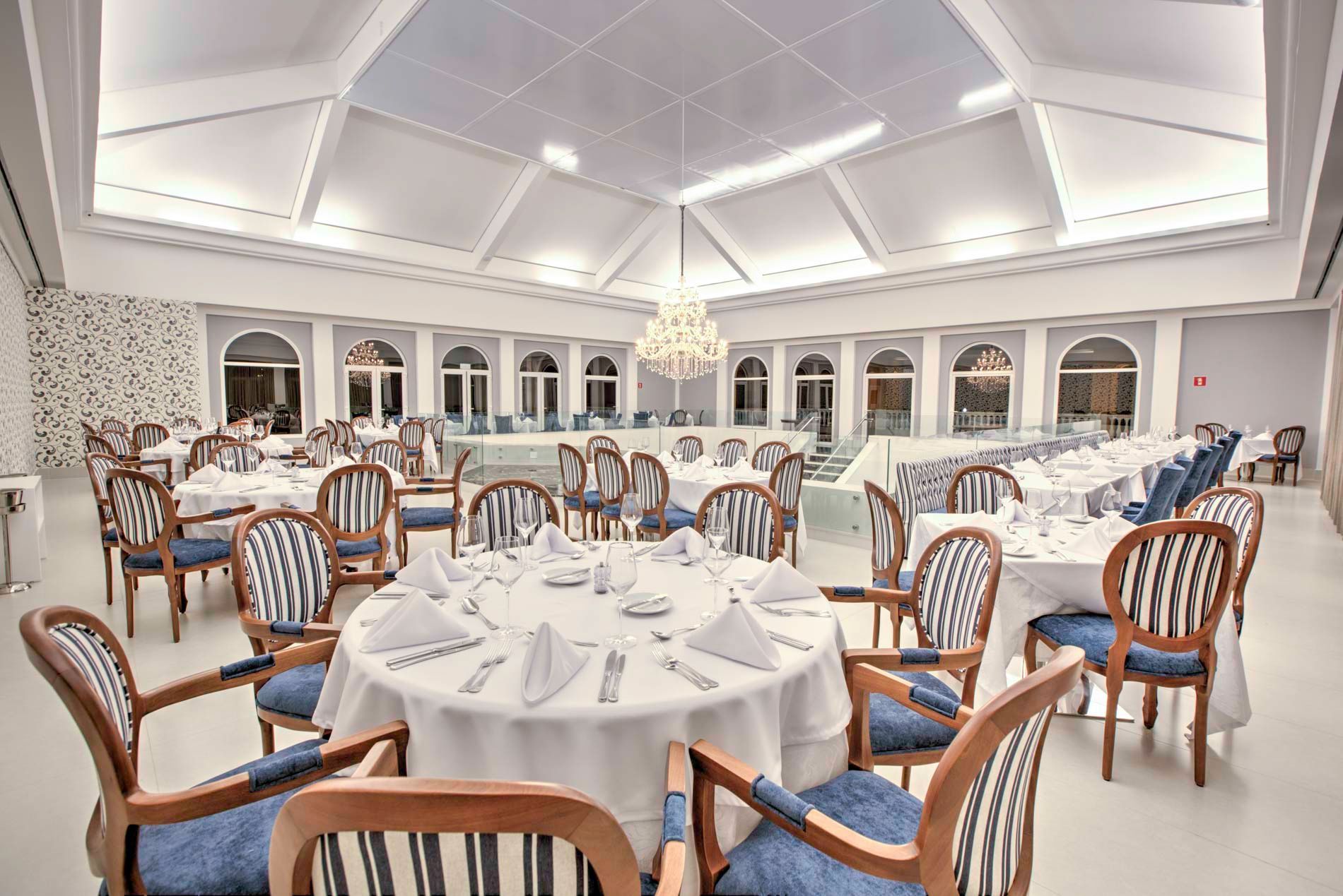 Elegante salão de jantar com mesas redondas preparadas para refeições. Cadeiras azuis e brancas, lustre e janelas em arco.