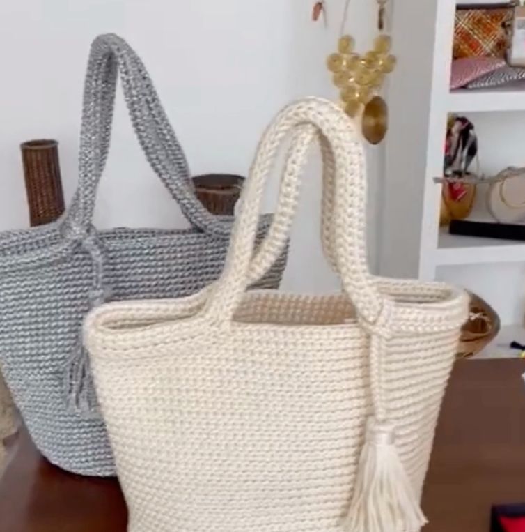 Duas bolsas de crochê, uma cinza, outra creme, com alças e borlas. Sobre uma mesa.