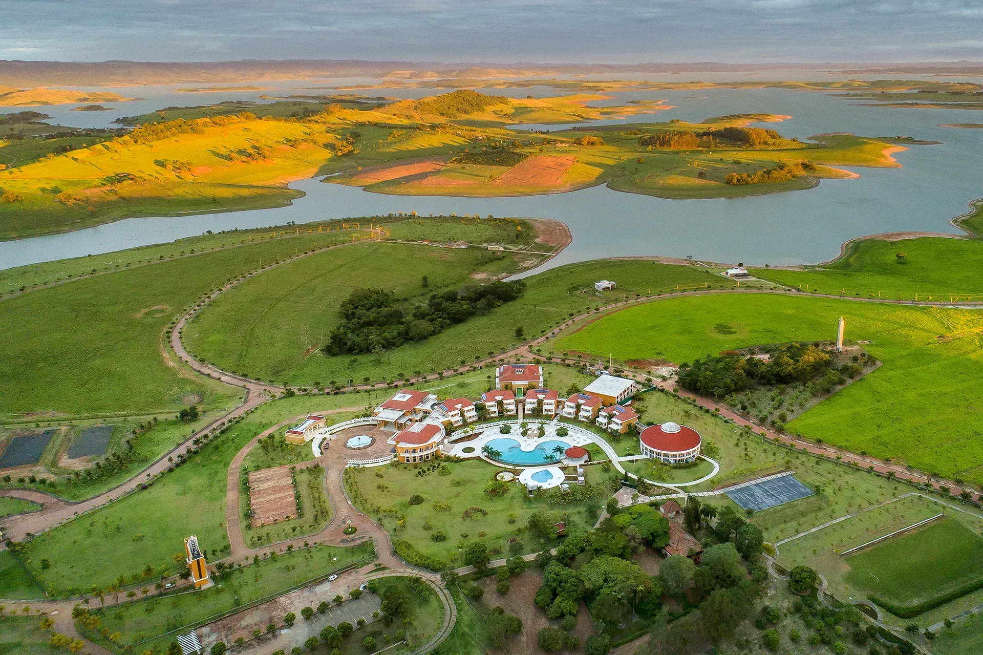 Vista aérea de um resort com piscina, cercado por campos verdes e um lago sob um céu nublado.