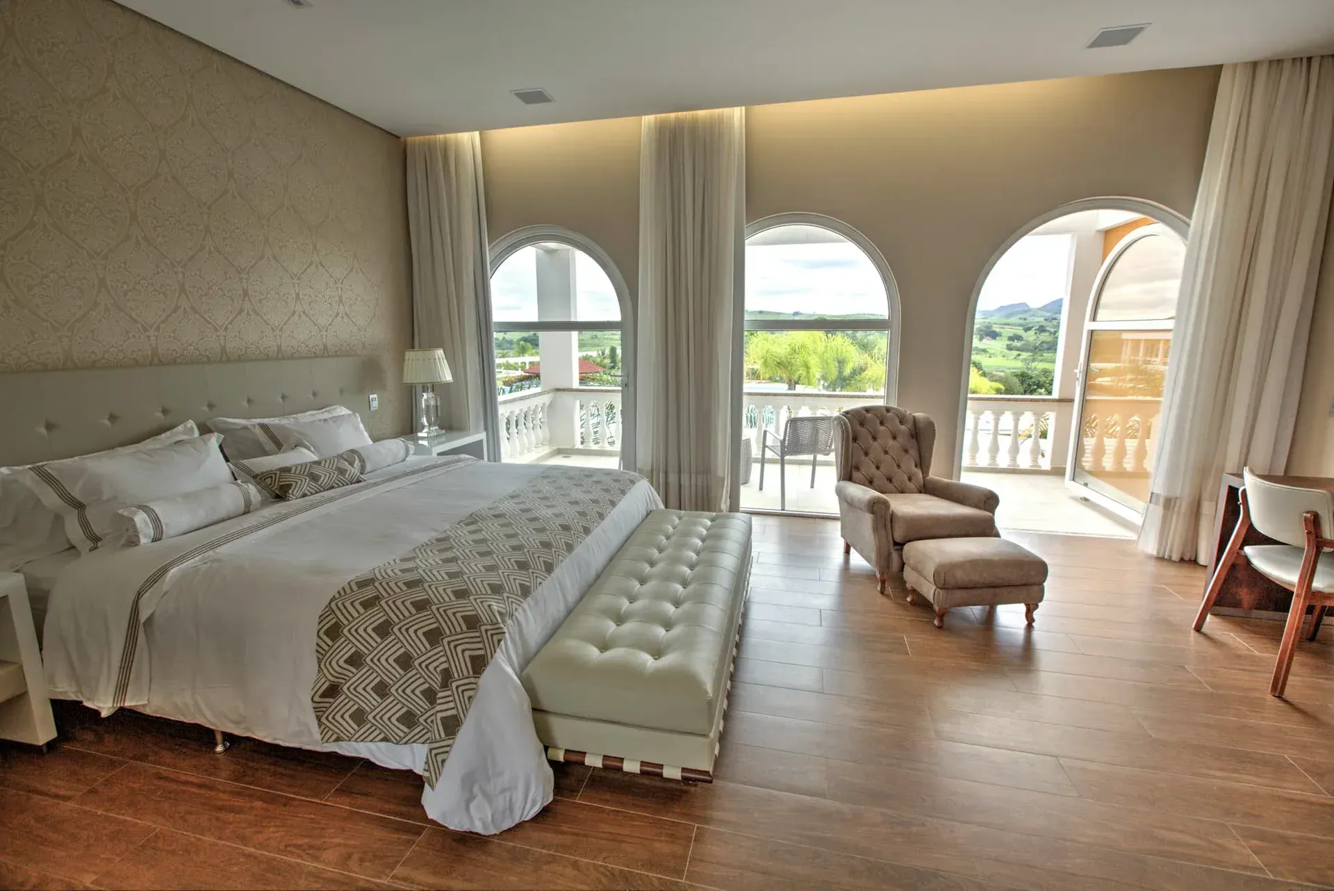 Quarto luxuoso com cama king size, janelas em arco, poltrona e acesso à varanda.