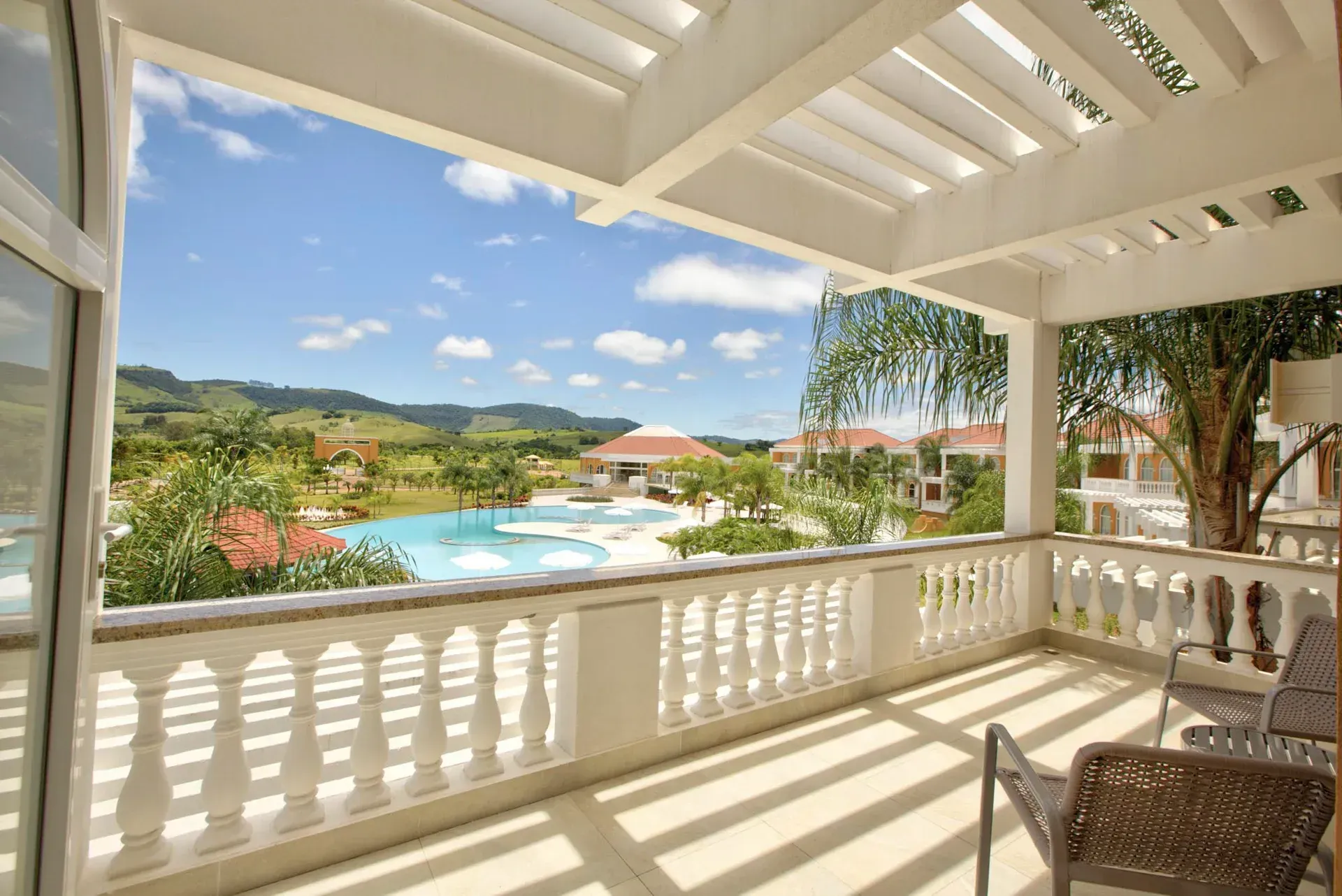 Varanda com vista para um resort com piscina, céu azul e montanhas. Duas cadeiras na varanda.