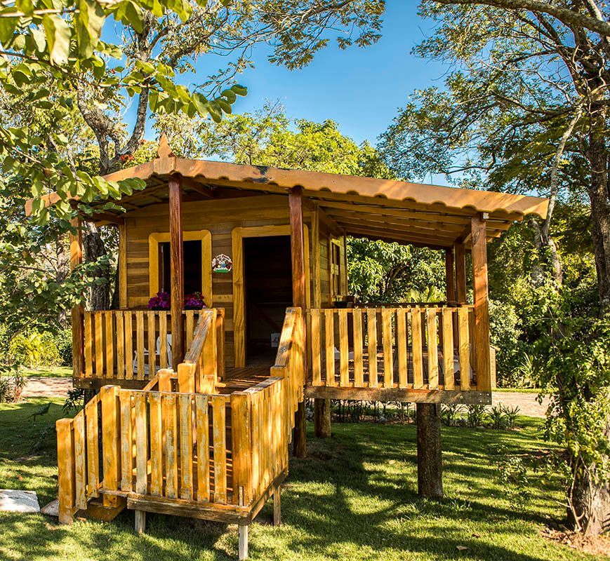 Casa na árvore de madeira com deck e grade, cercada por grama verde e árvores, sob um céu azul.