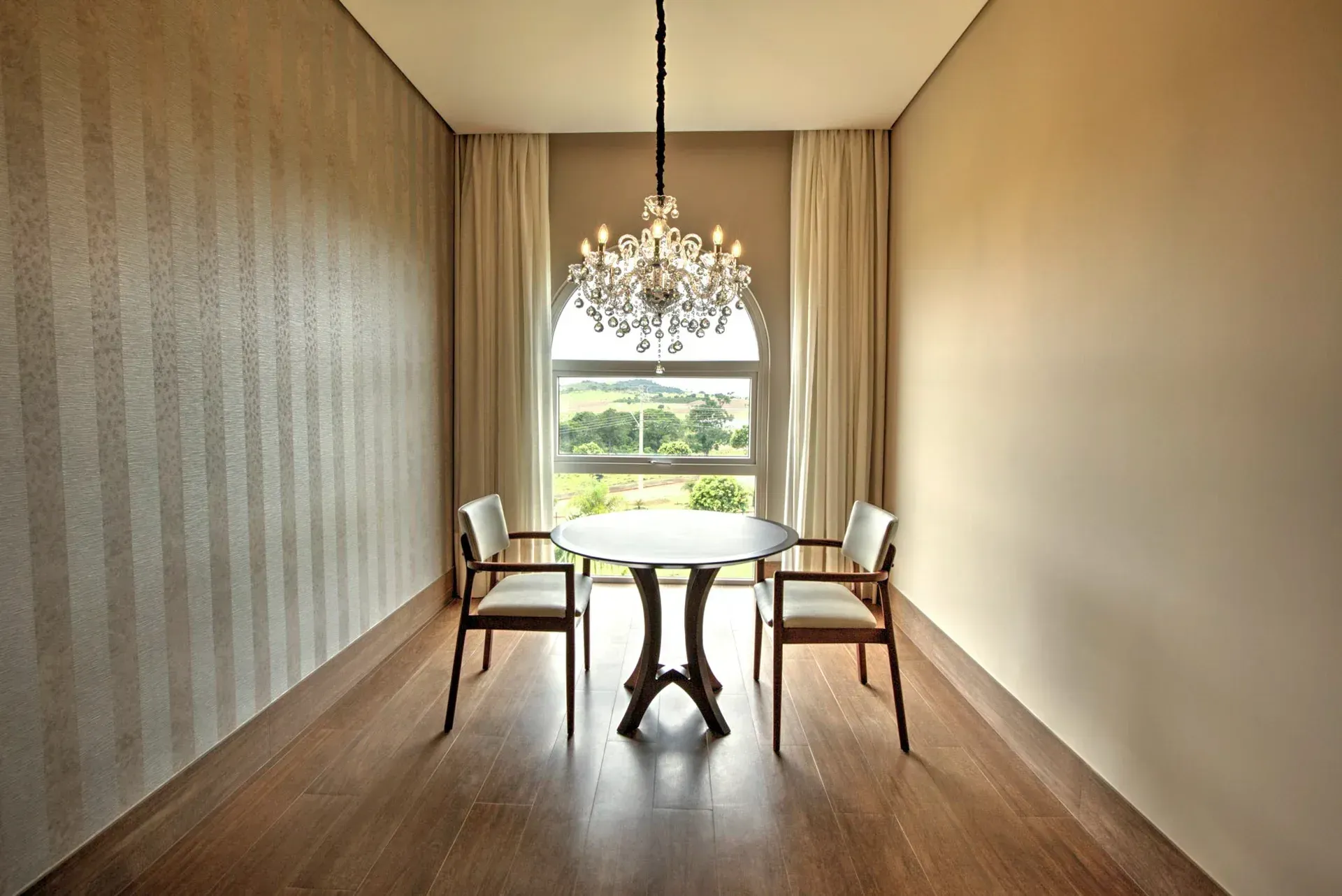 Sala de jantar com mesa redonda, duas cadeiras, janela, lustre e paredes bege.