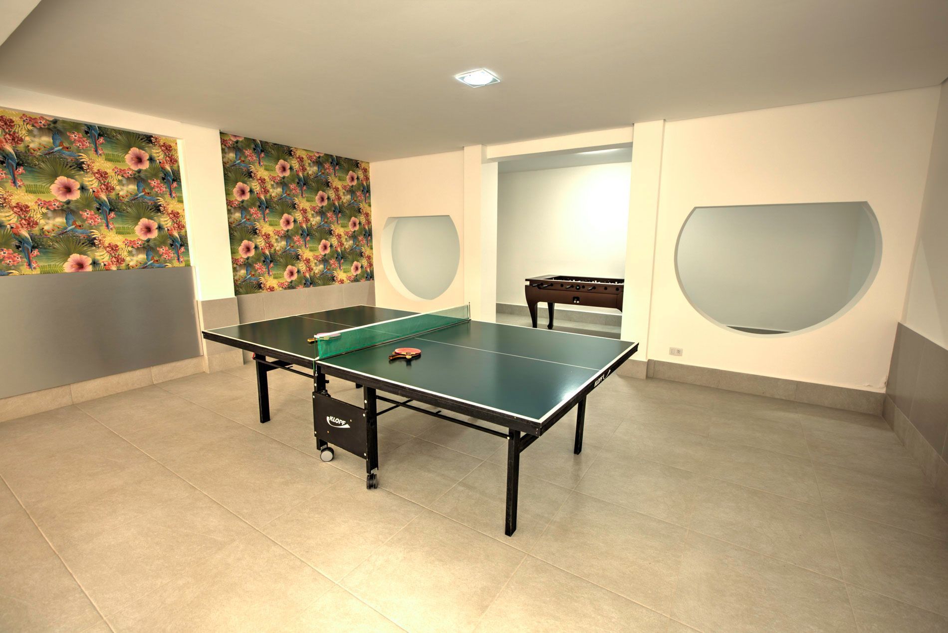 Mesa de pingue-pongue em uma sala bem iluminada com papel de parede floral e um vislumbre de uma mesa de sinuca ao longe.