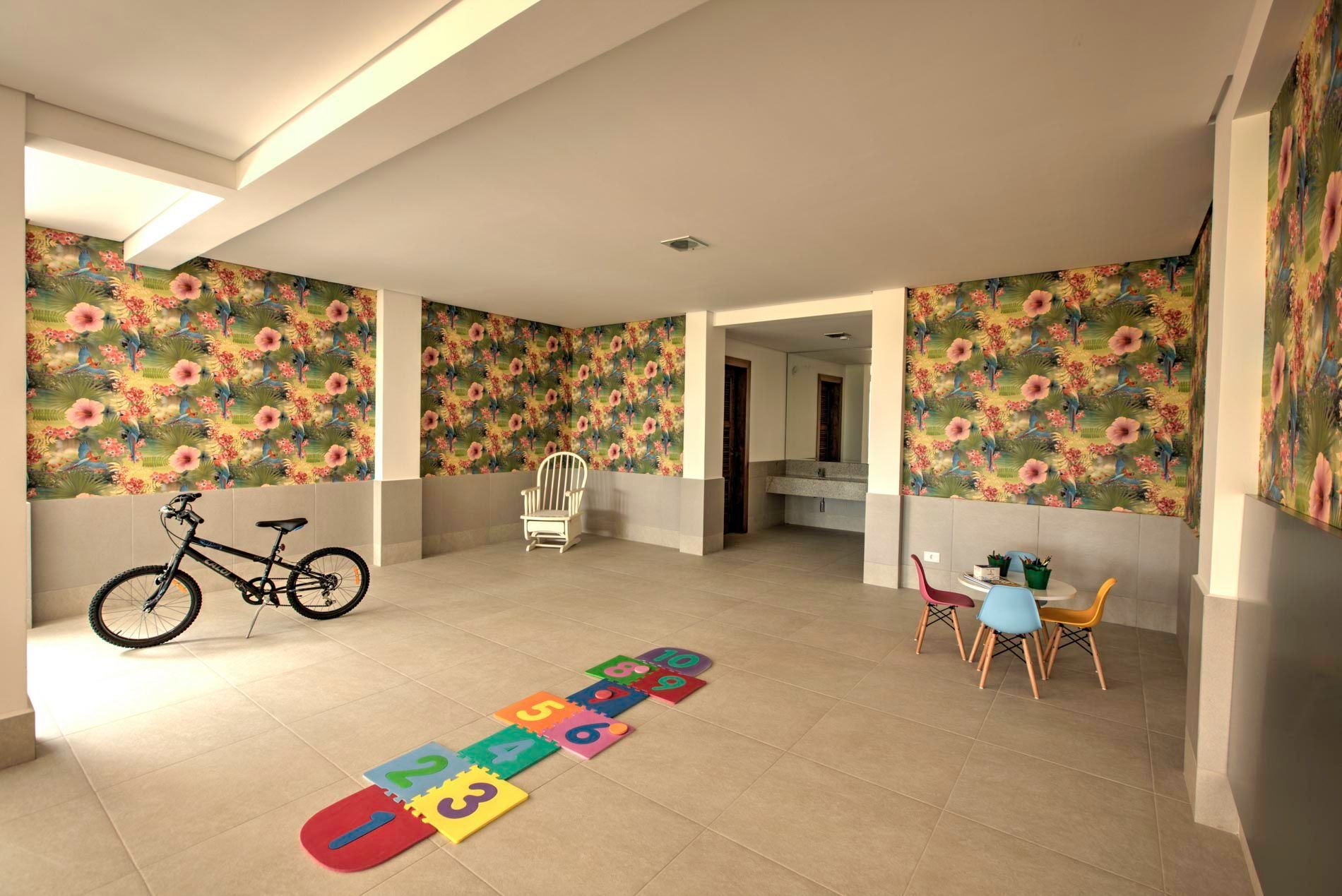 Sala de jogos com papel de parede floral, bicicleta, tapete de amarelinha e mesa pequena com cadeiras.