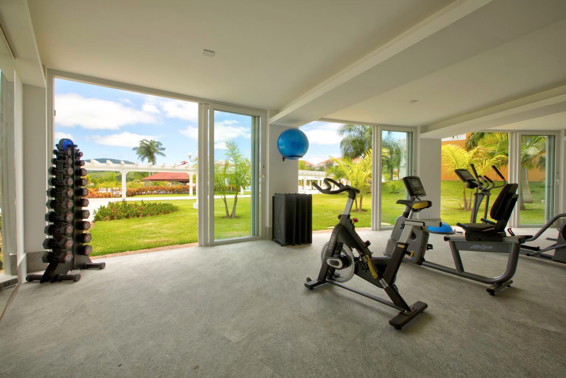 Academia com pesos, bicicletas ergométricas e bicicleta reclinada perto de janelas com vista para um jardim.