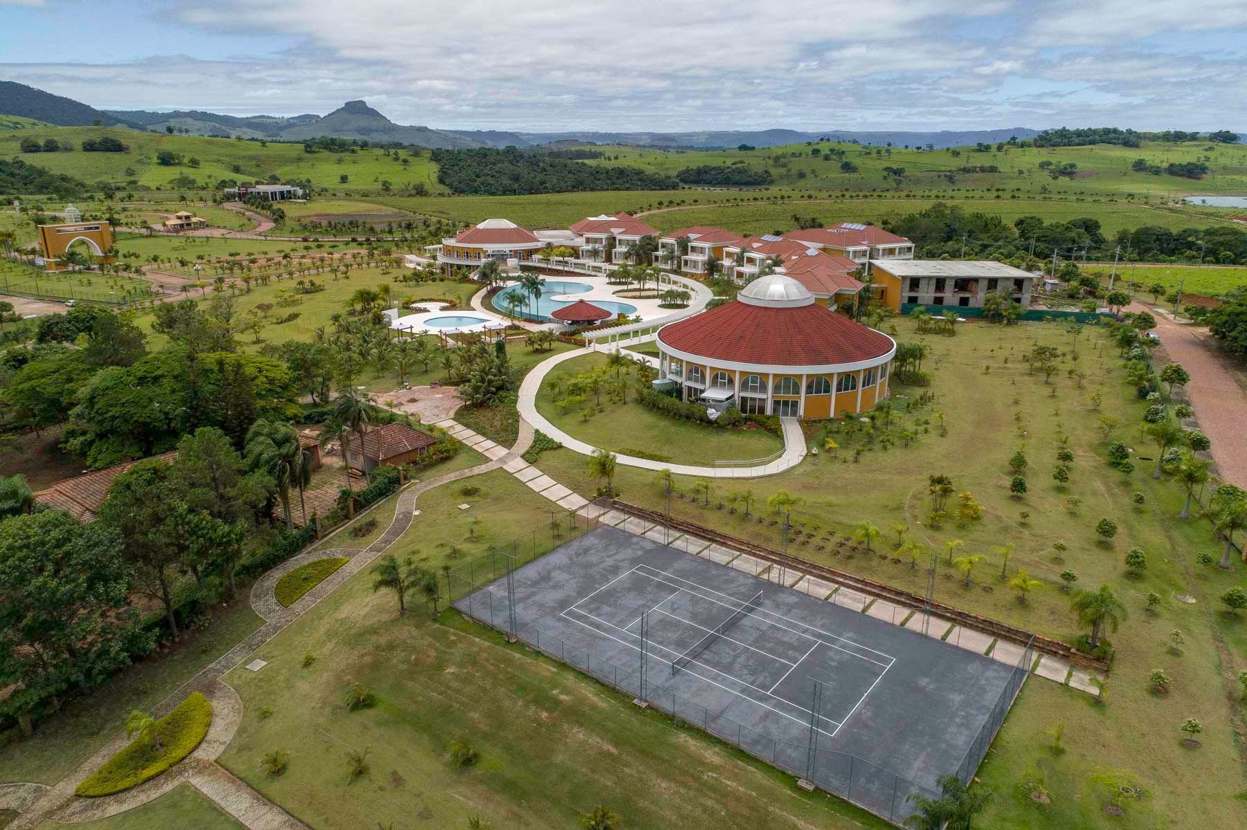 Vista aérea de um resort com piscina, quadra de tênis e edifícios cercados por campos verdes e colinas.