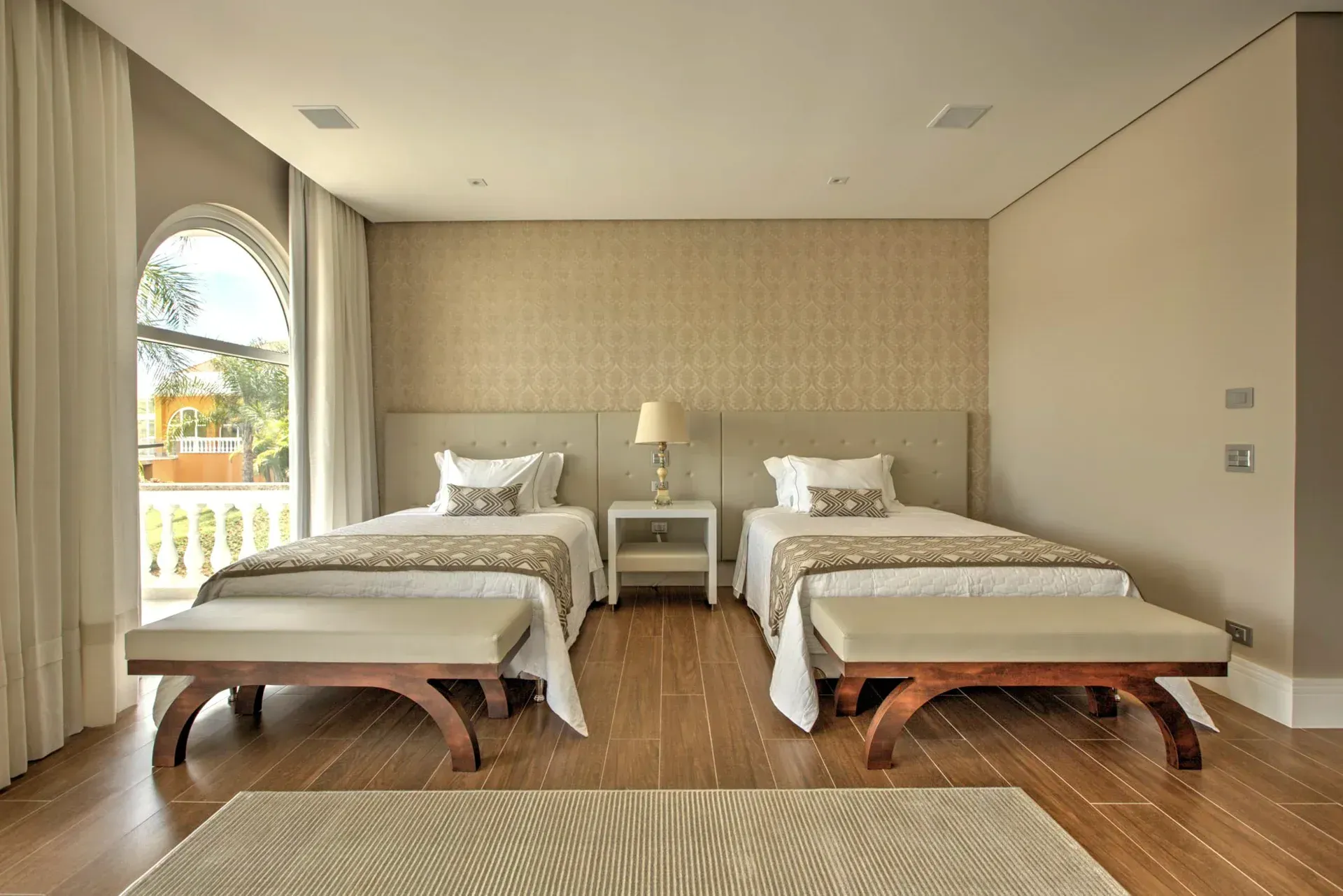 Duas camas de solteiro com roupa de cama bege e branca em um quarto com parede estampada e janela em arco.