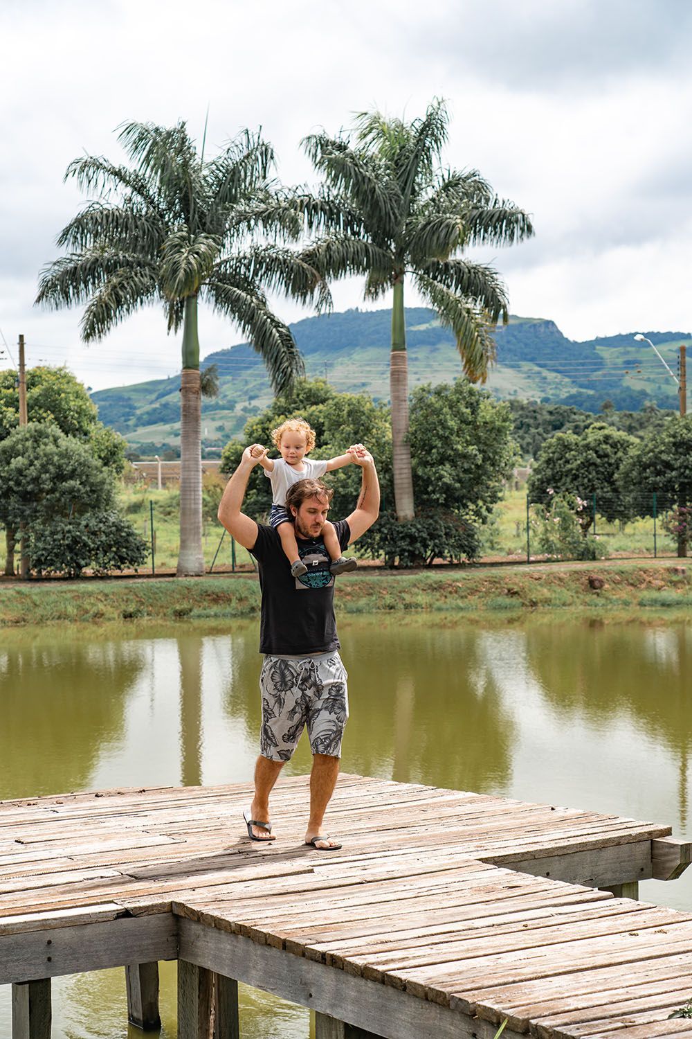Homem carregando criança nos ombros em um cais de madeira perto de um lago, com palmeiras e uma montanha ao fundo.