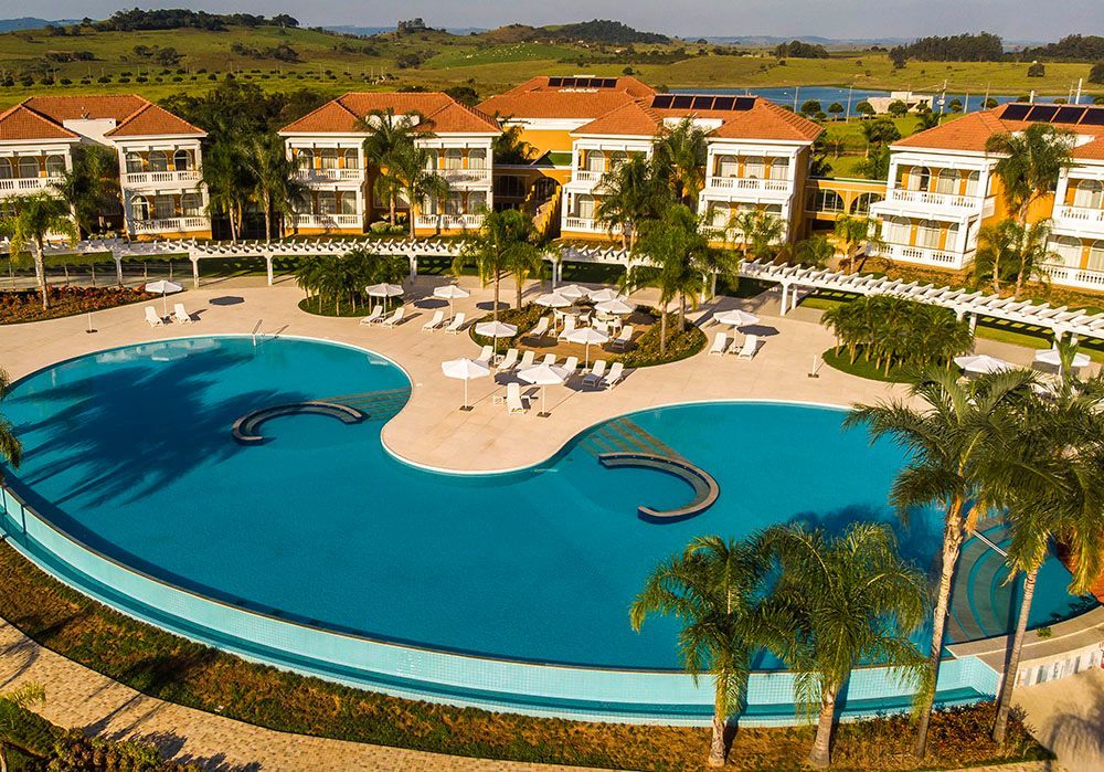 Resort com grande piscina azul, espreguiçadeiras, palmeiras e prédios de vários andares.