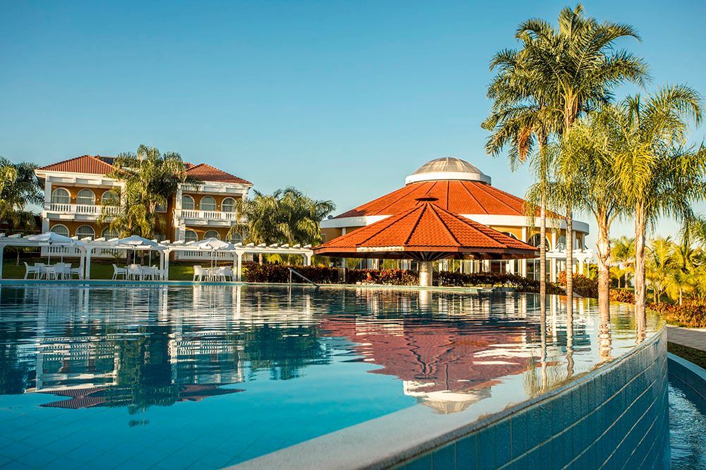 Piscina com reflexos, edifícios e palmeiras sob um céu azul brilhante.