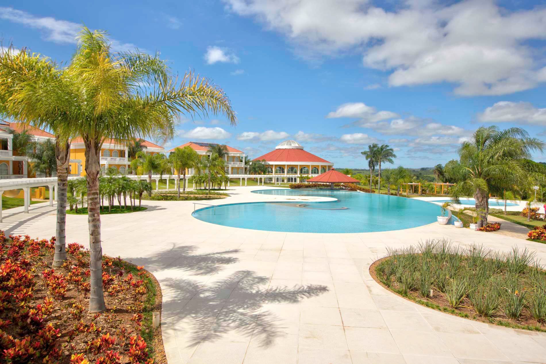 Resort tropical com piscina azul, palmeiras e céu ensolarado.