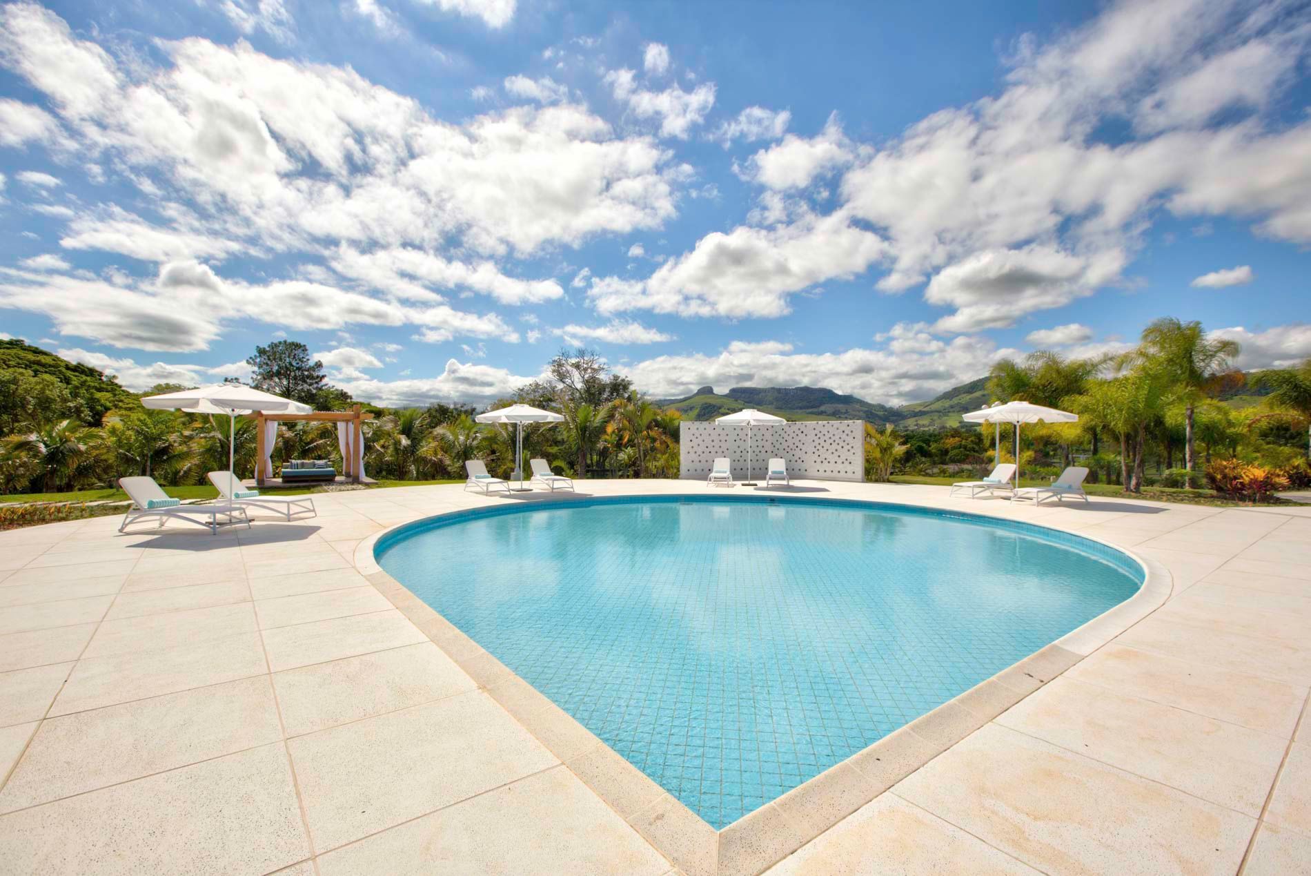 Piscina com água azul, espreguiçadeiras brancas e guarda-sóis sob um céu claro e nublado, cercada por vegetação.