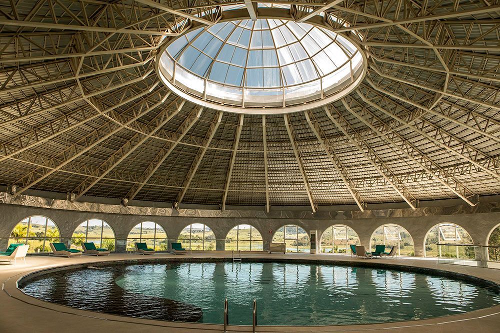 Piscina circular coberta sob uma cúpula com claraboia; espreguiçadeiras ao redor da piscina.