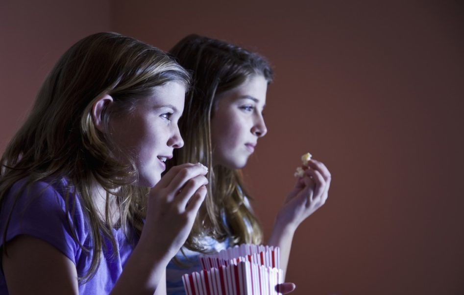 Duas meninas assistindo a um filme no escuro, comendo pipoca. Uma conversa, a outra segura pipoca.