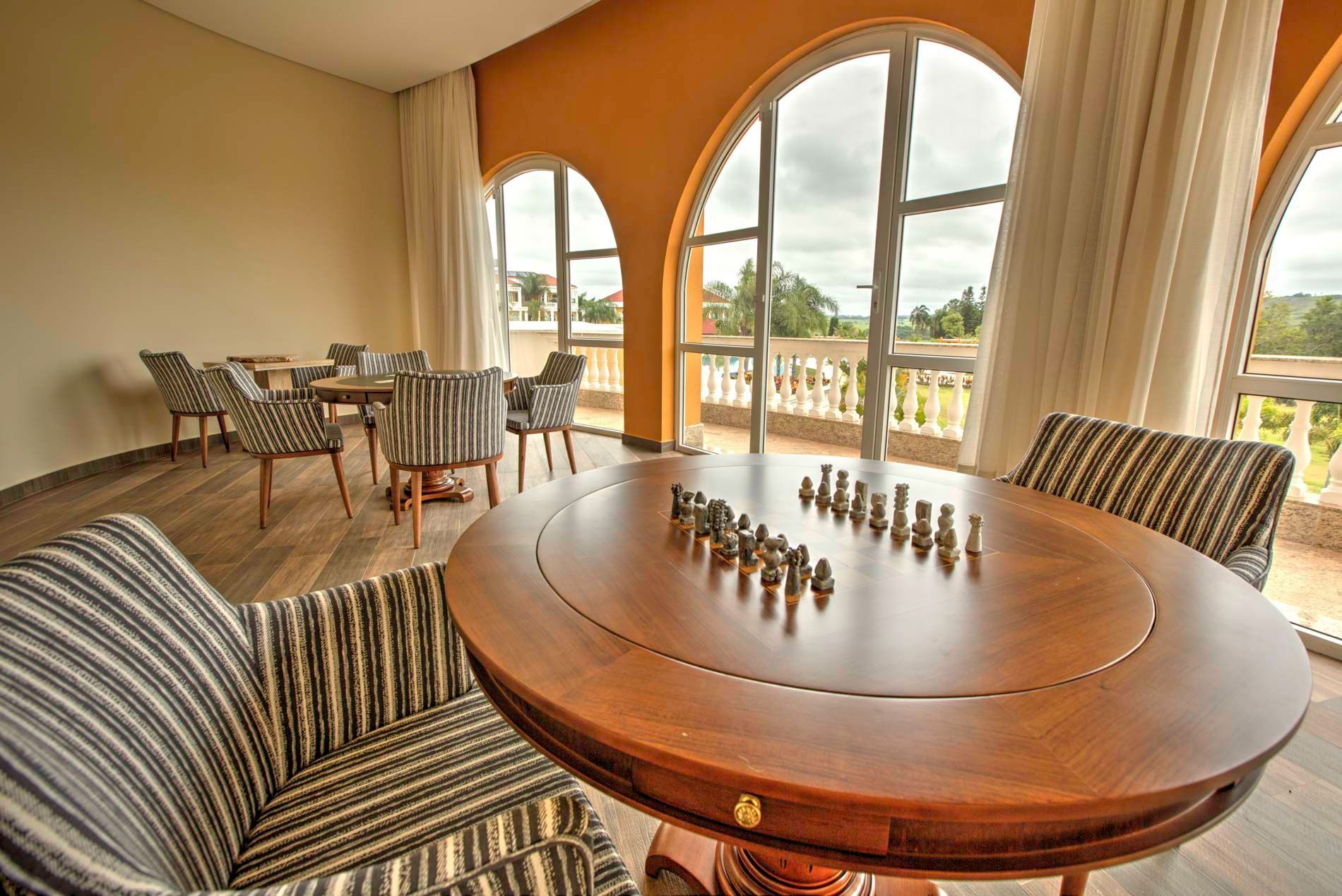 Tabuleiro de xadrez sobre uma mesa redonda, cercado por cadeiras listradas, em uma sala com janelas em arco